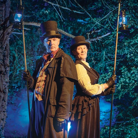 Two people in victorian costumes with lanterns at the edge of woodland