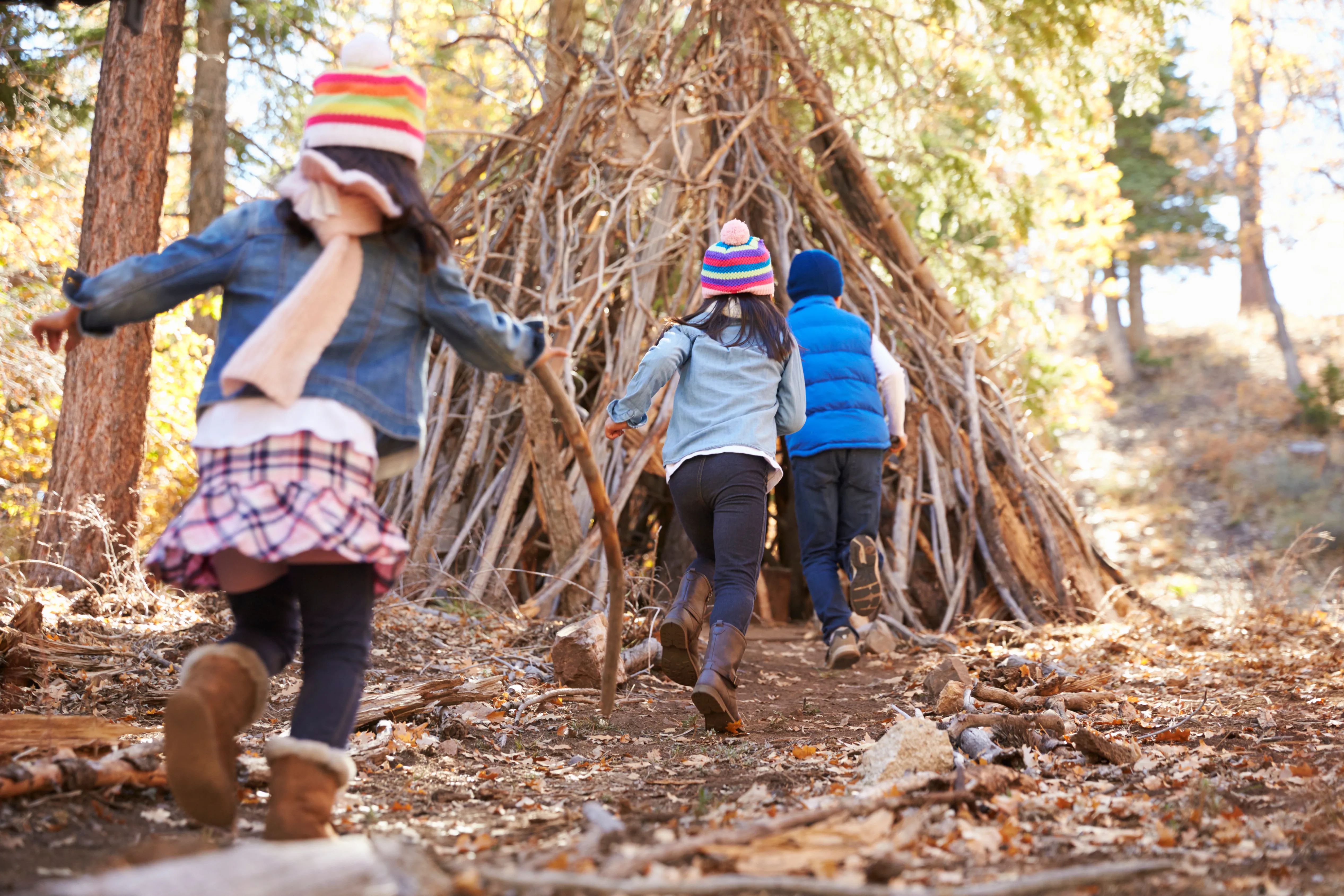 children running towards a den built of sticks in an autumnal woodland setting