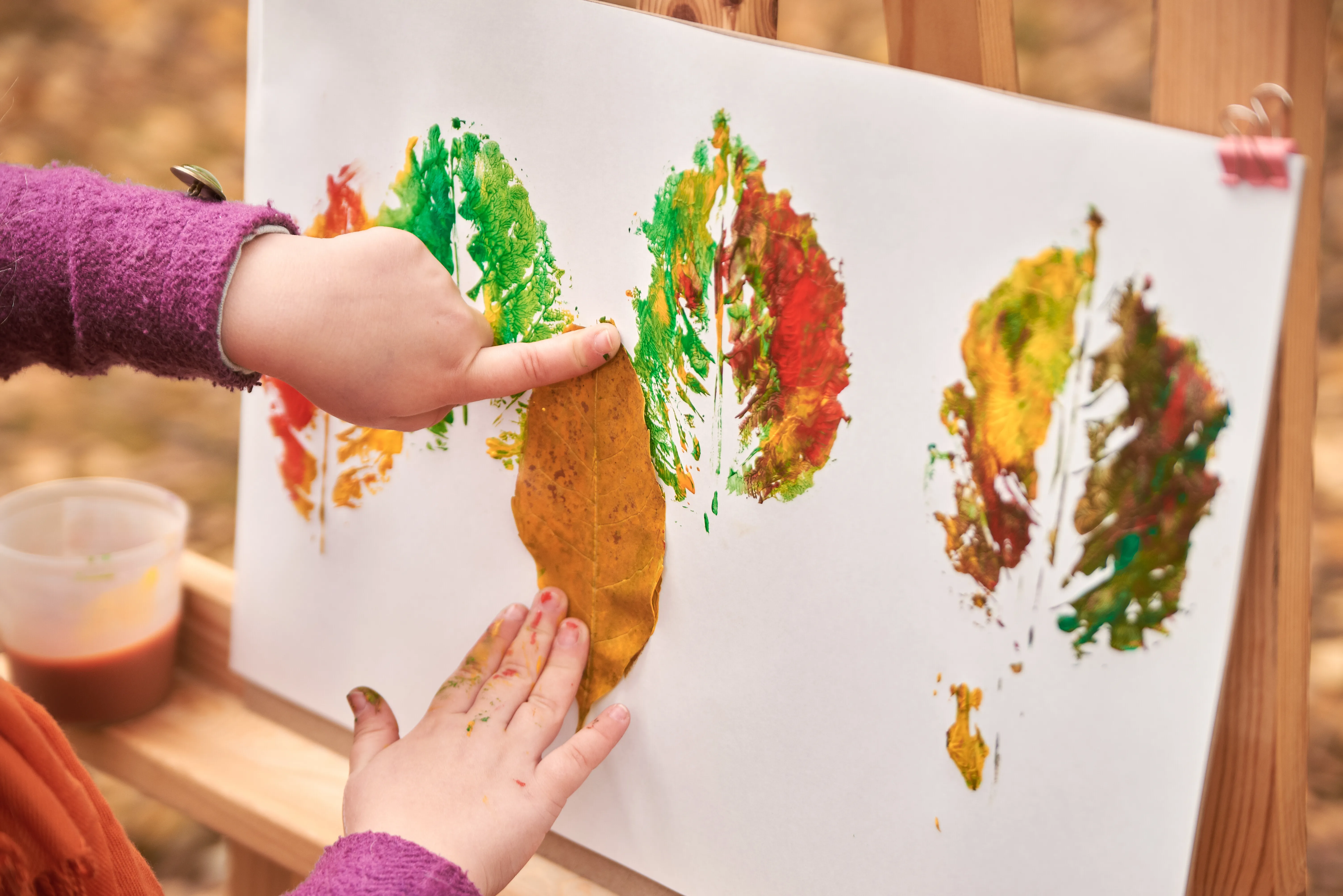 prints of autumn leaves on a wide paper, with a childs hand making a new print.