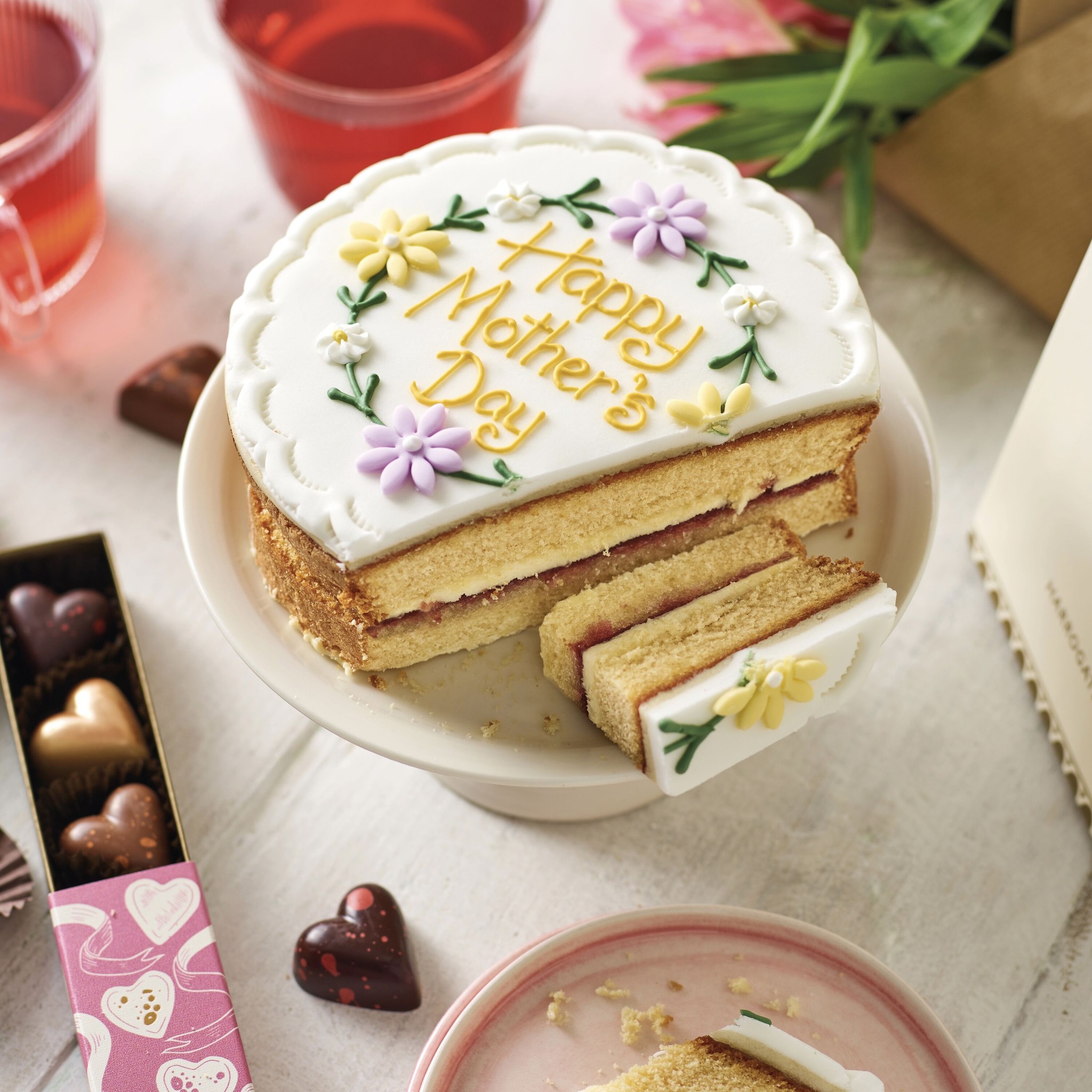 Betty's Victoria sponge cake sliced to show inside, iced with white icing, flowers and words Happy Mothers Day