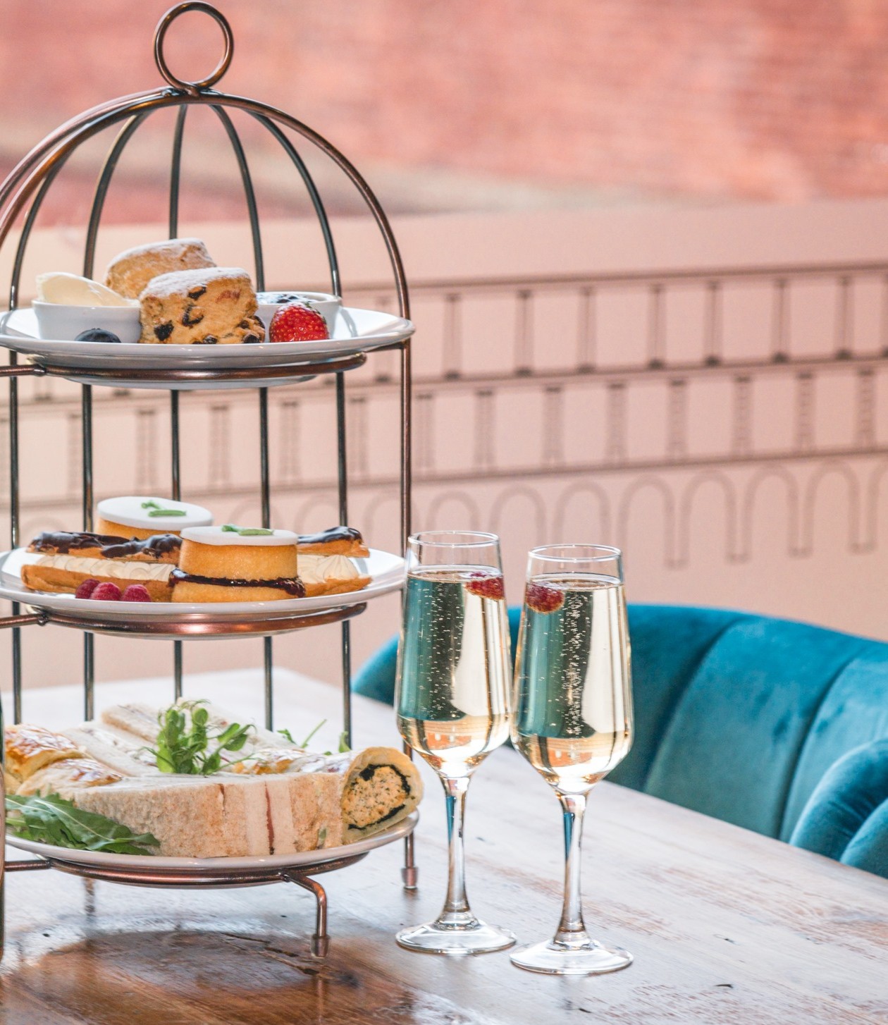 afternoon tea cakes and sandwiches presented on a three layer bird cage style platter, next to two glasses of prosecco