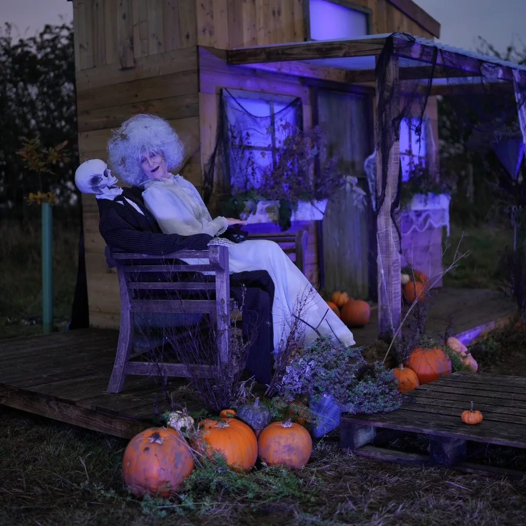 ghostly white figure sitting on a skeletons knee in a big chair, pumpkins are on the ground, ramshackle shack in the background